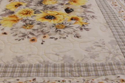 A close-up of a quilt with a floral pattern featuring large yellow and white flowers, surrounded by decorative borders and plaid accents in soft, neutral colors. The quilt is spread out on a flat surface.