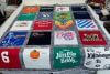 A colorful patchwork quilt made from various t-shirts with different logos, graphics, and text, laid out on a wooden bed frame in a cluttered room.