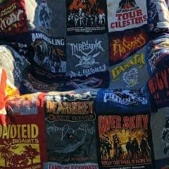 A quilt draped over a chair outdoors, made from various colorful rock band t-shirts featuring band names, logos, and album art. The background includes grass, trees, and a blurry house.