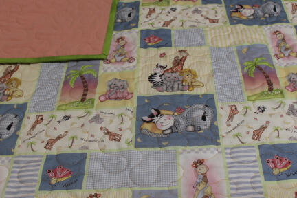 A colorful patchwork baby quilt with animal prints including giraffes, elephants, zebras, and lions on a wooden table. The quilt has a pink underside and green trim.