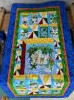 A colorful quilt with a central panel featuring cartoon jungle animals, bordered by blue, green, red, and yellow geometric patterns, draped over a wooden table in a craft room.