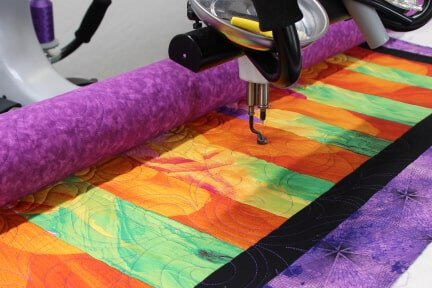 A quilting machine stitches a colorful quilt with orange, green, yellow, and purple fabric, bordered by black and purple edges. The machine’s needle is actively sewing intricate patterns on the quilt.