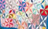 A colorful patchwork quilt with a floral geometric pattern and blue border is draped over a rustic wooden fence, with grass and trees in the background.