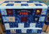 A handmade quilt featuring various colorful fabric squares, some with cartoon prints and others with embroidery like U.S. Forest Service FIRE and Mason 2023, draped over a table in a craft room.