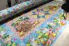 A colorful Easter-themed quilt is being stitched on a sewing machine, featuring patterns of eggs, bunnies, flowers, and a wooden cart filled with spring flowers on pastel backgrounds.