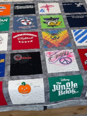 A colorful patchwork quilt made from various t-shirts with different logos, graphics, and text, laid out on a wooden bed frame in a cluttered room.