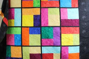 A colorful patchwork quilt with large squares and rectangles in bright colors such as red, yellow, blue, green, and purple, bordered by black strips, displayed in a craft room with shelves and drawers in the background.