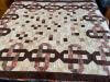 A handmade quilt featuring a pattern of interlocking brown, pink, and cream geometric shapes forming a chain-like design, with a swirled quilting pattern across the entire surface.