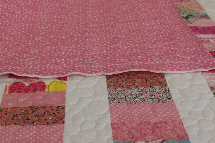 A colorful quilt with a white, textured border and patterned strips in pink, blue, and floral fabrics lies on a table. The top corner is folded back, revealing a pink backing with white polka dots.
