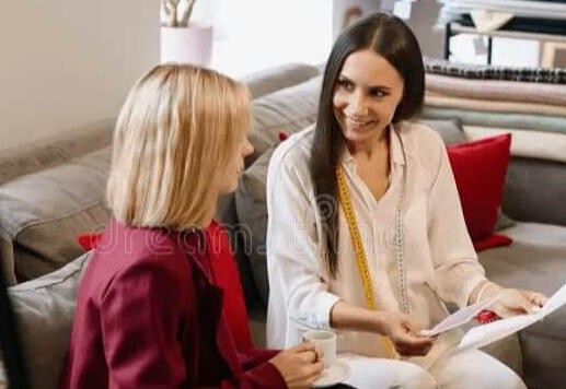 Two women sit on a couch, talking and holding papers. One wears a red jacket, the other a white outfit. They appear to be having a discussion over coffee in a cozy, modern room with fabric samples in the background.