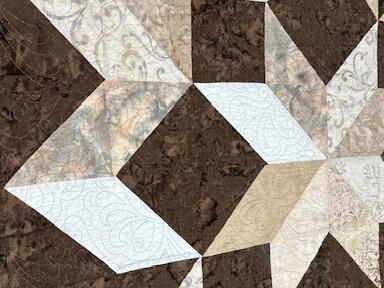 A close-up of a quilt featuring a geometric star pattern in shades of brown, cream, white, and beige, with intricate stitching visible throughout the design.