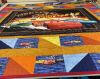 A colorful quilt featuring Disney Pixar Cars characters, including Lightning McQueen, is spread on a table surrounded by storage bins, quilting supplies, and sewing equipment in a craft room.