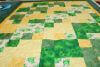 A colorful patchwork quilt with green, yellow, and white squares, some featuring playful cartoon frogs, is spread out on a flat surface.