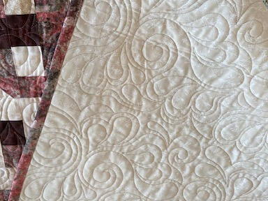 A handmade quilt with a patchwork pattern in earthy tones lies folded back, revealing intricate swirling quilted stitching on the cream underside.