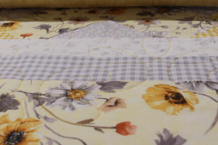 A close-up of a quilt featuring yellow and purple floral patterns, checkered sections, and intricate stitching, with a rolled quilt edge at the top.