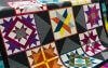 A quilt with colorful, geometric patchwork squares featuring star and diamond patterns in shades of purple, orange, blue, yellow, and green, bordered by dark gray triangles.