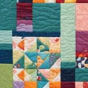 A colorful patchwork quilt featuring various geometric patterns and shapes in vibrant shades of blue, green, orange, red, pink, and purple, with visible stitching throughout.