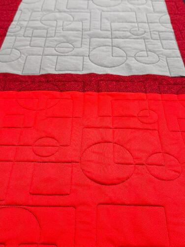 A close-up of a quilt made from various colored fabric squares, including red, gray, black, and striped patterns, stitched with geometric quilt patterns such as circles and squares. Sweatshirts made into a memory quilt