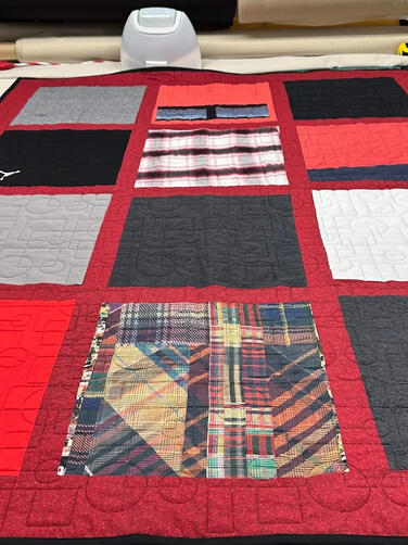 A handmade quilt with red borders and various patterned and solid fabric squares, including plaid and gray, laid out on a worktable in a sewing or crafting room. Causal Sports shirts made into Memory quilt