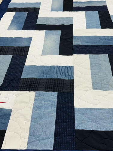 A handmade quilt featuring blue, white, and black rectangular patches in a geometric pattern, with a red Nike logo stitched on a corner. The quilt is displayed on a worktable with sewing tools in the background. Demin jeans memory quilt