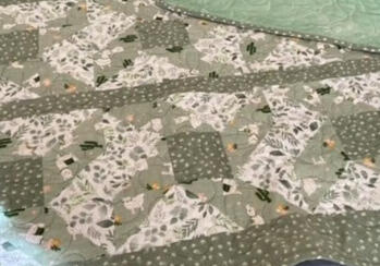 A handmade quilt with green and white patterned fabric, featuring cactus and nature motifs, lies on a bed. The quilt has a green polka dot border and a light green backing with stitched designs.