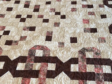 A handmade quilt featuring a pattern of interlocking brown, pink, and cream geometric shapes and squares on a cream background, with swirled quilt stitching visible across the surface.